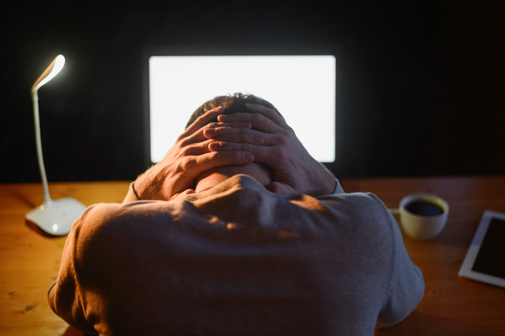 Homem de frente ao computador e com a mão na nuca e cabeça pra baixo em sinal de tristeza.