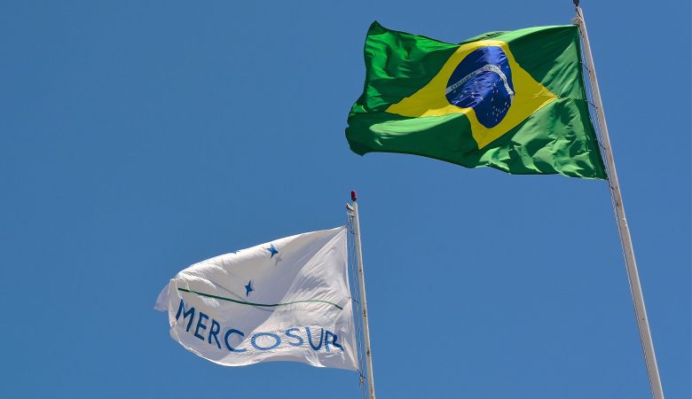 Foto das bandeiras do Mercosul e Brasil no poste tremulando.